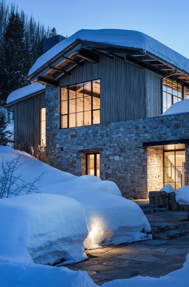 Exterior view of a multi-level mountain-style home heavily covered in snow. The building combines rough natural stone walls, vertical timber cladding, exposed timber eaves and dark metal-framed floor-to-ceiling glazing. Warm interior lighting contrasts with the cool blue evening light and snowy landscape. A stone-paved path leads toward the entrance.