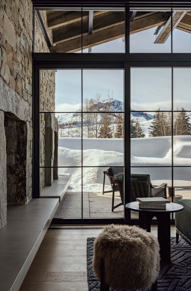 A refined mountain living area with expansive black-framed floor-to-ceiling glazing overlooking a snow-covered landscape. The interior features a substantial rough-stone wall and fireplace surround, exposed timber roof beams visible above the glazing, pale timber flooring, a dark geometric rug, leather lounge chairs, a dark sofa, and a furry ottoman around a small round coffee table. The space has a warm, rustic-luxe alpine character with strong indoor-outdoor connection.