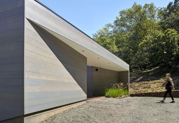 A minimalist contemporary single-storey building set into a wooded sloping site. The structure has a long, low horizontal form with smooth pale grey horizontal cladding, a deeply recessed covered entry or terrace, sharp angular wall geometry, and a flat roof with recessed downlights. Gravel paving and native grasses surround the entrance, while mature trees form the backdrop.