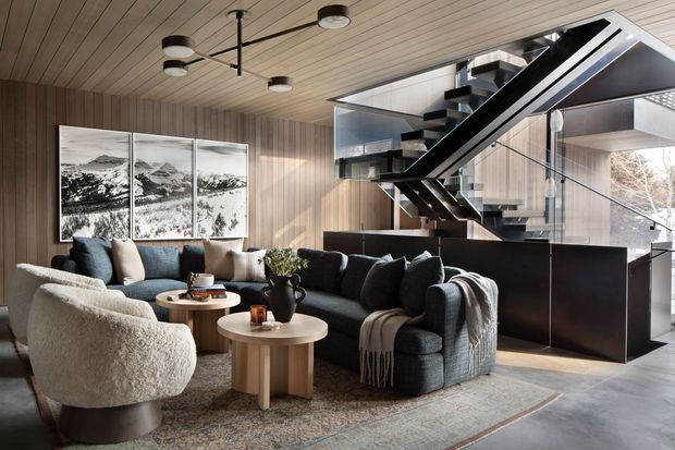 A refined open-plan living room with a large charcoal sectional sofa, sculptural boucle armchair, layered coffee tables, and a patterned area rug. Vertical timber wall panelling and a matching timber-lined ceiling create warmth, while a dramatic black open-riser staircase with glass balustrades dominates the background. Large glazing brings in natural light, and contemporary black-and-white artwork and a sculptural ceiling light complete the space.