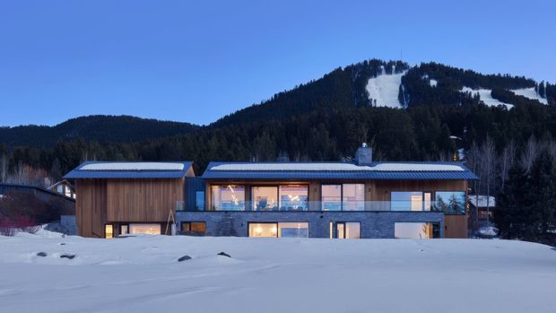 Wide exterior view of a contemporary mountain residence set in a snow-covered landscape. The long, low building has timber-clad sections, extensive glazing, a dark grey lower level, and shallow pitched metal roofs. A forested, snow-covered ski slope rises behind the house at dusk.