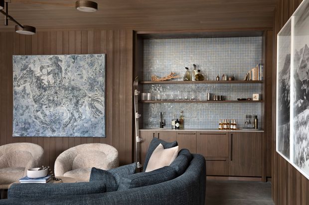 A sophisticated living room or lounge with a deep charcoal-blue curved sofa in the foreground and two rounded upholstered armchairs. The room features warm vertical timber wall panelling and a matching timber ceiling, dark timber built-in bar cabinetry with open shelving, and a small blue-grey mosaic tile backsplash. Contemporary artwork, sculptural lighting, glassware, and bottles add a refined, intimate atmosphere. A large framed artwork is also visible through reflection or glazing at the right edge.