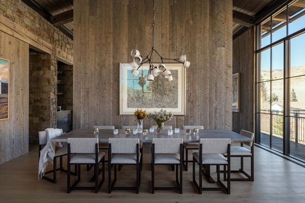 A sophisticated dining area with a long rectangular dining table and seating for approximately ten people. The room features a dramatic double-height timber-clad wall, exposed dark timber ceiling structure, rugged natural stone walls, a sculptural multi-light pendant, and expansive black-framed glazing opening to a balcony or exterior view. Neutral upholstered chairs, a large framed artwork, and floral table arrangements create a refined yet warm atmosphere.