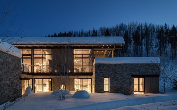 Snow-covered contemporary mountain residence at dusk, composed of dark stone-clad volumes framing a warmly illuminated glazed central living space. The building has a low-slope roof, extensive timber-framed glazing, exposed structural elements, and a sheltered upper-level terrace or balcony, set against a wooded snowy hillside.
