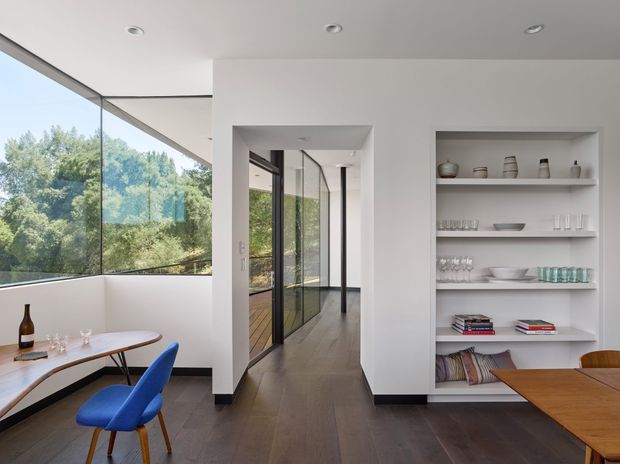 A bright, contemporary dining area with floor-to-ceiling corner glazing overlooking leafy trees. The room features white walls, dark timber flooring, a built-in open shelving unit displaying ceramics, glassware and books, and a light timber dining table with vivid blue upholstered chairs. A glazed corridor extends through the centre of the space.