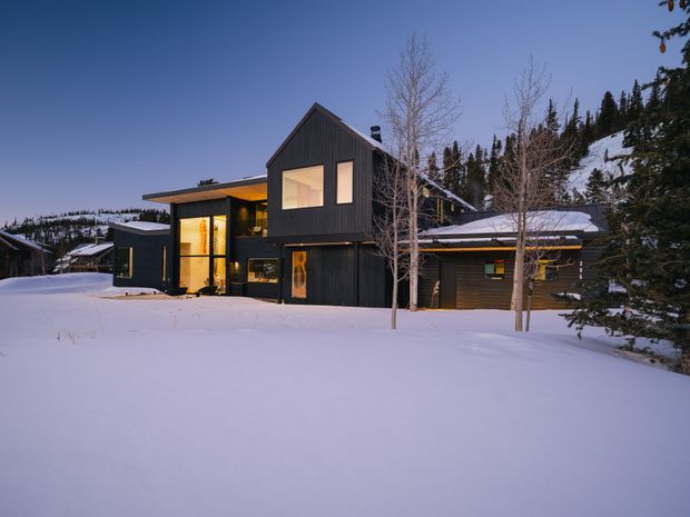 Exterior view of a contemporary multi-level house in a snowy alpine setting at dusk. The home features dark vertical cladding, a steep gabled central volume, broad roof overhangs, warm illuminated glazing, and connected low-profile wings. Snow blankets the foreground and surrounding landscape, with coniferous trees and wooded hills in the background.