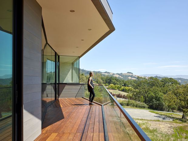 A contemporary upper-level balcony or terrace extends along a modern home, with warm timber decking, frameless or minimally framed glass balustrades, extensive floor-to-ceiling glazing, and a deep cantilevered roof soffit with recessed downlights. The terrace overlooks a leafy, rolling landscape under a clear sky.