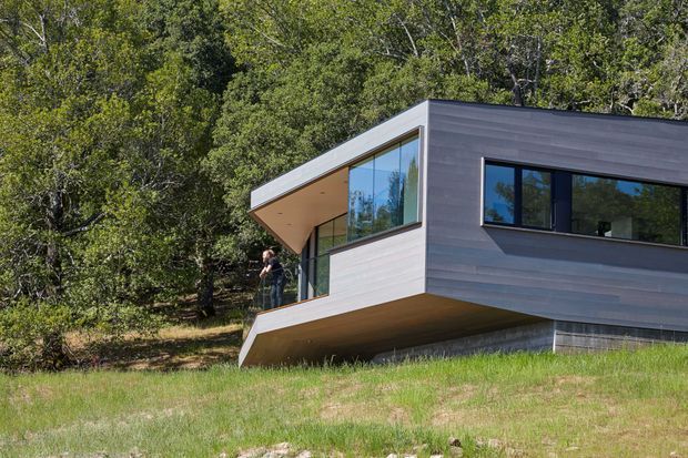 Exterior view of a modern single-storey house set into a wooded, sloping landscape. The building has a sharply angled roofline, cantilevered upper volume, extensive corner glazing, dark horizontal cladding, and a partly recessed lower level. The composition emphasises a strong connection to the surrounding trees and hillside.