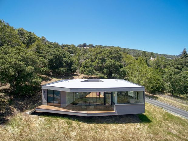 A compact contemporary single-storey home set among wooded rolling hills. The building has a broad low-pitched or nearly flat roof with a central roof opening, extensive floor-to-ceiling glazing, dark exterior framing and cladding, and a projecting timber-toned deck along the front. A narrow road passes beside the house, with dense vegetation and dry grass surrounding the site.