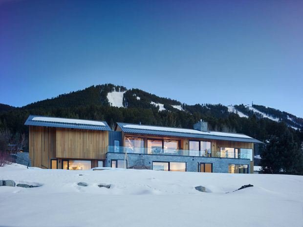 A contemporary multi-level mountain house photographed in winter, set against a forested, snow-covered alpine slope. The building combines dark stone or masonry lower walls, extensive glazing, and vertical timber-clad upper volumes beneath low-pitched metal roofs. Warm interior lighting is visible through the windows and balconies.