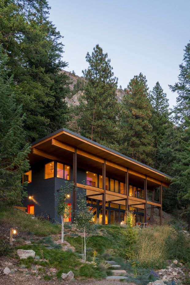 A contemporary two-storey mountain house nestled among dense pine forest and rocky terrain. The home features a long low-pitched roof with deep timber soffits, dark horizontal exterior cladding, extensive warm-lit glazing, timber posts and beams, and an elevated covered balcony or veranda. A stone stepping path and landscaped native grasses lead toward the house, with a rugged mountain slope visible behind the trees.