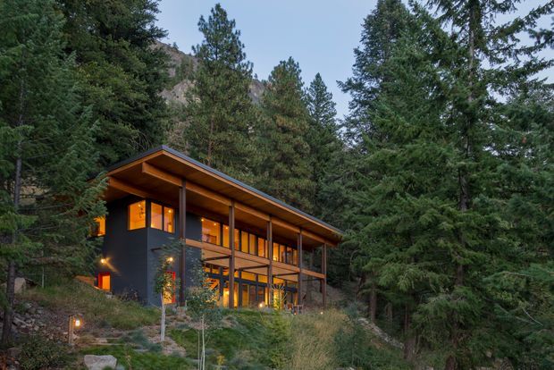A modern two-storey mountain home nestled among dense evergreen trees on a steep wooded site. The dark-clad building has extensive warm-lit glazing, slender vertical structural posts, and a broad projecting single-slope roof with exposed timber soffit. Rocky terrain and a mountain slope are visible behind the house, creating a secluded forest setting.