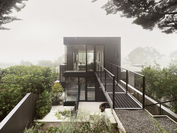 A modern multi-level house set among mature trees and dense landscaping, viewed from an elevated exterior walkway. The compact rectilinear building has dark-framed floor-to-ceiling glazing, a flat roof, dark solid wall panels, and a narrow metal-grate bridge with black railings leading to the upper level. Lower-level glazing and a landscaped path are visible below, creating a strong elevated, cantilevered composition integrated into the sloping wooded site.