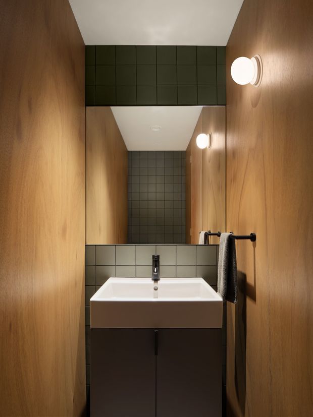 A compact, narrow bathroom viewed directly toward a wall-mounted rectangular white basin with dark vanity cabinetry below. Warm timber wall panels line both sides, while dark square tiles form the backsplash and rear wall. A large mirror creates a reflected view down the space, complemented by round wall lights and a black towel rail. The design is restrained, contemporary, and softly illuminated.
