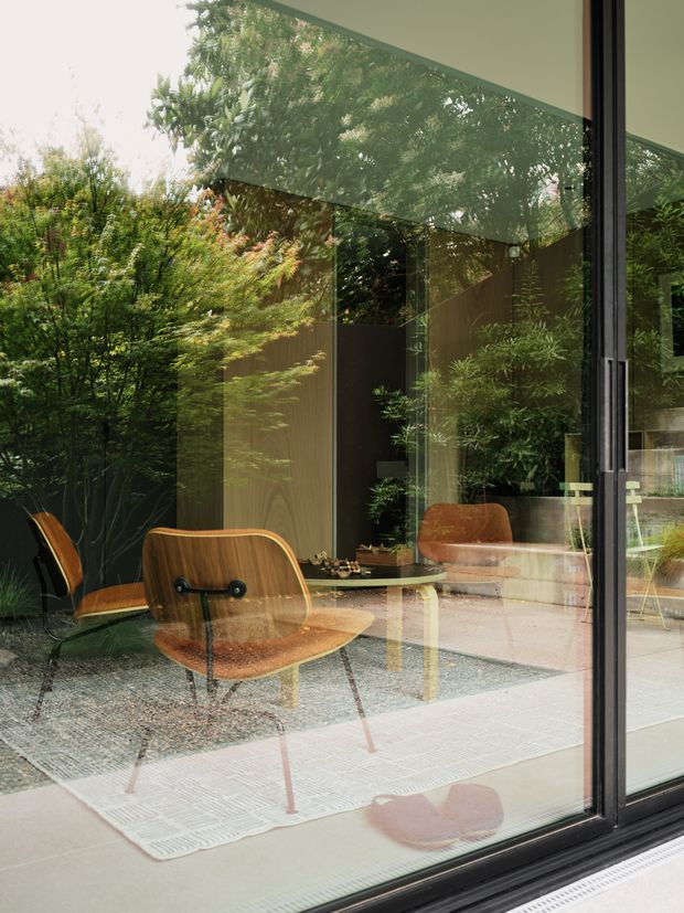 A contemporary living room viewed through expansive floor-to-ceiling glazing, with mid-century-inspired wooden lounge chairs, a low coffee table, textured area rug, and dark built-in wall surfaces. The glazing strongly reflects and reveals a lush planted courtyard or garden, blending the interior with the greenery outside. Black-framed sliding or folding glass doors define the edge of the room.