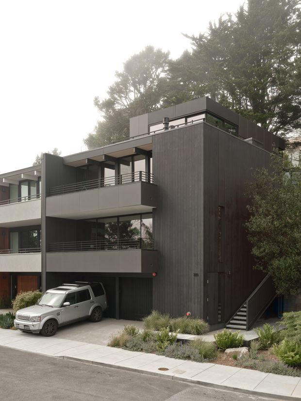 Exterior view of a contemporary multi-storey urban residence with a dark vertical-cladding feature volume, flat rooflines, recessed balconies, horizontal metal balustrades, large glazed openings, and an integrated ground-level garage. A landscaped frontage with ornamental grasses and shrubs borders the concrete driveway and footpath.