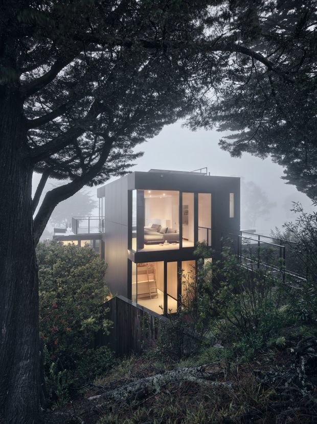 A moody exterior view of a compact contemporary multi-level house set among dense mature trees on a steep, vegetated site. The dark, box-like building has extensive floor-to-ceiling glazing, warm illuminated interiors, projecting balconies or terraces with slim metal railings, and a flat roof. A large tree frames the foreground, while misty conditions create a secluded forested atmosphere.