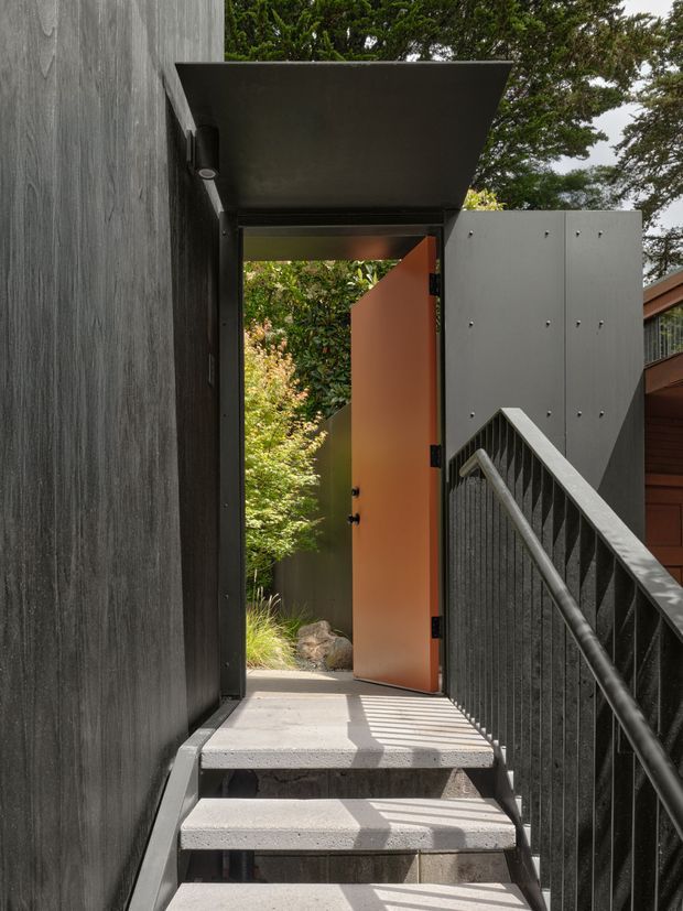 A narrow exterior stairway leads to an open doorway in a dark, textured-clad building. The entrance features a vivid terracotta-orange door beneath a projecting flat canopy, with black metal railings and a perforated dark screen along the stairs. Dense landscaping is visible beyond the doorway, and part of an adjacent timber-clad structure appears at right.
