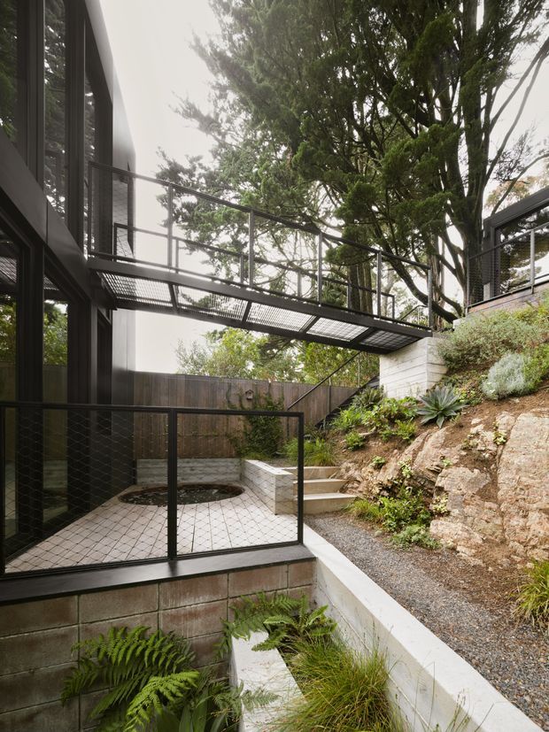 A contemporary multi-level residential exterior set into a landscaped sloping site. Dark-framed glazing and cladding frame a raised metal pedestrian bridge with mesh balustrades, linking the building across an open courtyard. Below is a paved terrace with a circular recessed feature, concrete block retaining walls, steps, gravel paths, exposed rock, and dense mature vegetation including ferns and succulents.