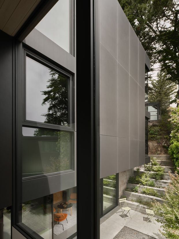 Close exterior view of a contemporary multi-level residence, focused on dark-framed floor-to-ceiling glazing and smooth light grey panel cladding. The glazing reveals a partial interior with orange seating, while landscaped stepped garden terraces and a concrete courtyard are visible beside the building. The composition emphasizes the facade, vertical window detailing, and connection to the planted site.