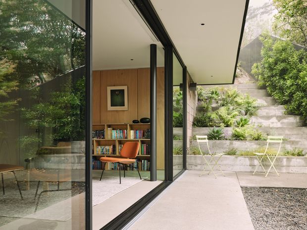 A contemporary indoor-outdoor living space viewed from a paved courtyard. Full-height black-framed glazing opens to a compact library or study with warm timber wall panelling, built-in bookshelves, artwork, and an orange lounge chair. A broad flat soffit extends over the exterior walkway, while stepped concrete garden beds with lush planting rise behind a small outdoor café table and chairs. Reflections in the glazing add depth to the scene.