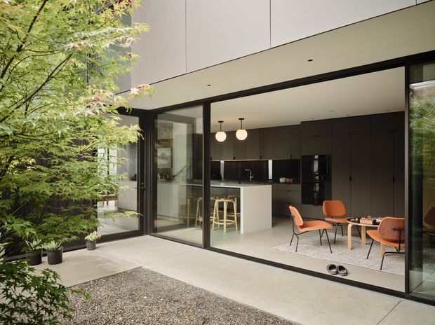 View from a landscaped courtyard into a contemporary open-plan kitchen and dining area through expansive black-framed glazing. The space features dark full-height kitchen cabinetry, a white island, pendant lights, polished pale flooring, and orange mid-century-inspired dining chairs. A gravel courtyard, concrete paving, and Japanese maple planting frame the exterior threshold.