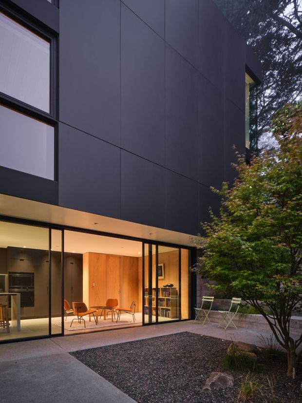 A contemporary two-storey house viewed from a landscaped courtyard at dusk. The facade is clad in large dark charcoal panels with black-framed glazing, while the lower level opens to a warmly lit interior featuring timber joinery, shelving, and mid-century-style seating. Concrete paving, a gravel garden bed, and planted trees define the outdoor space.