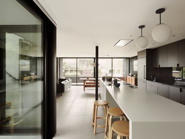 Open-plan contemporary kitchen and living area viewed from beside a large glazed opening. A long pale island with timber stools dominates the foreground, with dark built-in cabinetry, integrated appliances, a black splashback, pendant globe lights, tiled flooring, and full-height glazing opening toward an outdoor view. The living room is visible beyond the island.