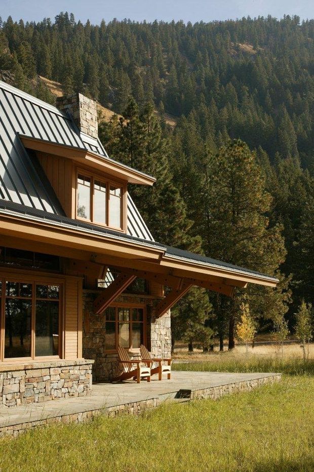 Exterior view of a rustic mountain lodge or cabin with a steep standing-seam metal roof, timber siding and exposed timber structure, stone chimney and masonry base, large timber-framed windows, and a broad covered terrace. The house sits beside a forested, mountainous landscape with a dry grass meadow.