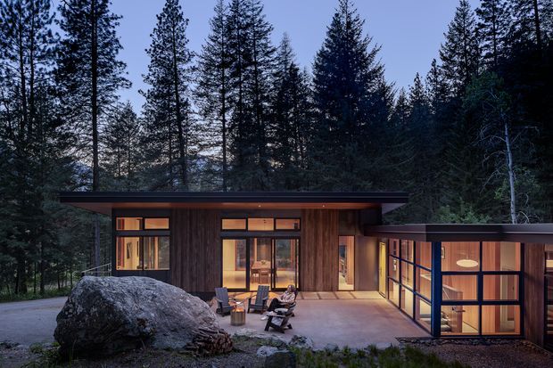 A low, modern single-storey cabin or residence nestled among tall conifer trees. The building features dark timber cladding, extensive floor-to-ceiling glazing, and broad flat roof planes, with a warm, softly illuminated interior visible through the windows. A large natural boulder and outdoor seating occupy the foreground, reinforcing the connection to the forested landscape.