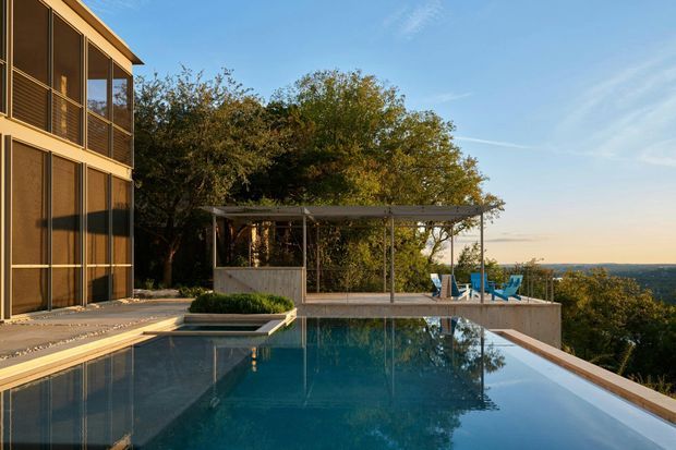 Exterior architectural photograph of a modern multi-level home beside an infinity-edge swimming pool. The building has extensive floor-to-ceiling glazing with dark frames, warm timber-toned exterior elements, and a flat roofline. A covered outdoor pavilion with a lightweight pergola structure sits alongside the pool, with landscaped trees and a distant coastal or ocean-like horizon beyond.