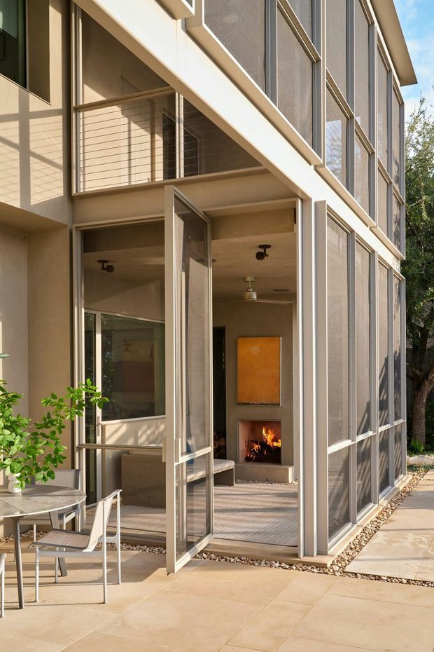 A contemporary multi-level residential exterior with a glazed indoor-outdoor living space. Large operable screened glass panels and open doors form a light-filled enclosure around a sitting area and fireplace, while a small outdoor dining setting occupies the paved terrace. The facade uses warm neutral render, slender light-coloured frames, extensive mesh-screened glazing, and strong horizontal and vertical geometric lines.