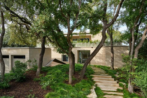 A low, modern residential building set within a mature, densely planted garden. The facade uses pale beige horizontal masonry or stone panels, with recessed dark-framed openings and large glazed doors. A stepped stone pathway rises through the landscaping toward the entrance, while an upper-level terrace with a planted or balustraded edge is visible behind the trees.