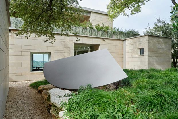 Exterior view of a low-profile contemporary building composed of pale rectangular stone or concrete-clad volumes. A planted roof edge and extensive grasses integrate the structure into a landscaped site, while a large abstract pale sculpture sits in the foreground beside a gravel path. Dark-framed windows and a recessed glazed opening provide limited views into the interior.
