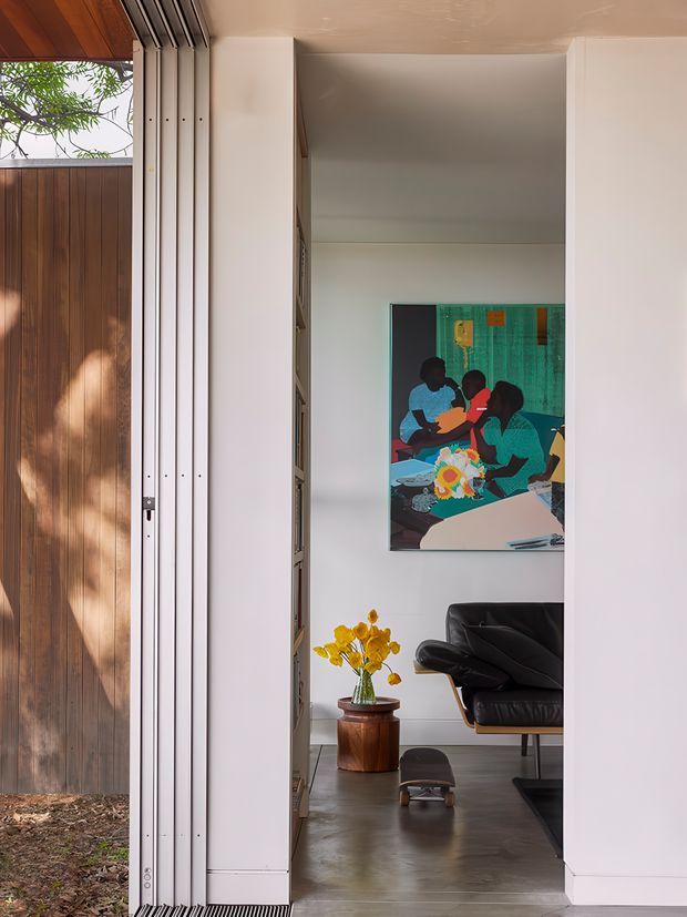 A view through a large open folding glass door into a refined living room. The space features white walls, a smooth grey concrete-like floor, a black leather lounge chair, a timber side table with yellow flowers, built-in shelving, and colourful wall artwork. A warm timber-clad exterior wall and landscaped courtyard are visible outside. The opening has slim light-coloured metal frames and a timber-lined soffit.