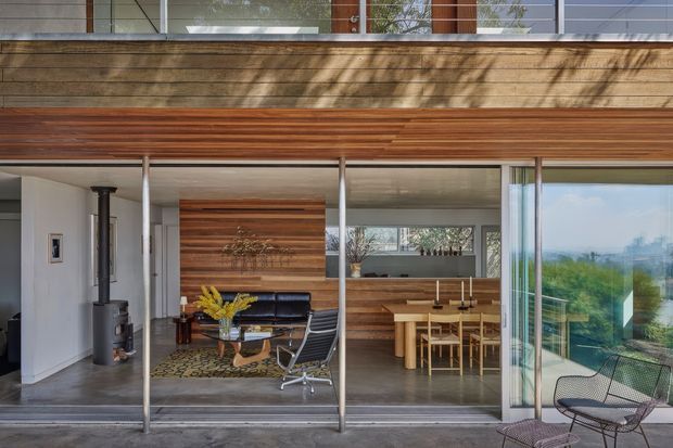 Exterior-facing view of a modern multi-level residence with extensive floor-to-ceiling glazing and a broad horizontal timber-clad facade. Through the open glazing, a combined living and dining area is visible, featuring timber wall panelling, a black wood-burning stove, dark leather seating, a dining table, and contemporary furnishings. The property overlooks a leafy landscape and distant urban skyline. The composition emphasizes indoor-outdoor connection, horizontal lines, natural timber, and transparent glass.