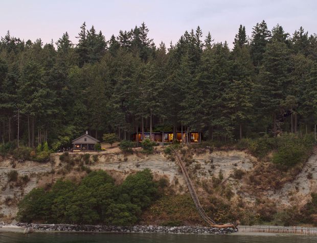 A distant view of a small cabin-like building nestled among dense evergreen forest on a steep, sandy, rocky waterfront slope. A narrow rail or track descends from the building toward a shoreline walkway and calm water. The structure is partially obscured by trees, with warm interior lighting visible through its windows.