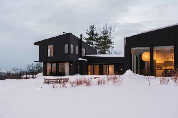 A modern dark-clad residential building set in a snowy landscape. The composition shows several connected cubic volumes with flat or very low-pitched roofs, large floor-to-ceiling glazed openings, warm interior lighting, and a partially enclosed outdoor terrace or living area. Snow-covered landscaping and leafless trees surround the house.