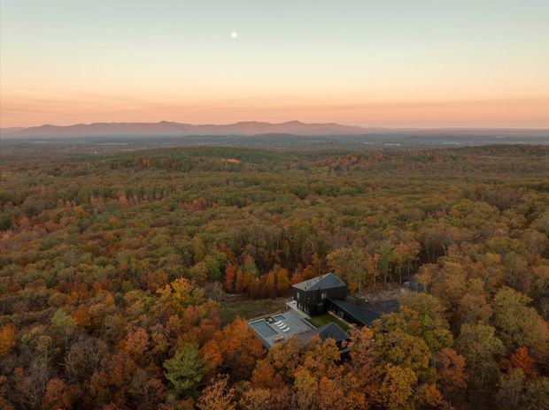 Aerial view of a dark-roofed, modern house nestled within dense autumn-coloured forest. The expansive wooded landscape stretches toward distant low mountain ridges beneath a pastel sunset sky, with the home partially visible in the lower foreground.