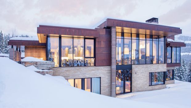 A contemporary multi-level mountain residence set in deep snow, with broad floor-to-ceiling glazing, projecting flat roof planes, warm-toned metal or timber cladding, and light masonry walls. Interior lighting glows through the windows, while snowy forested hills form the surrounding setting.