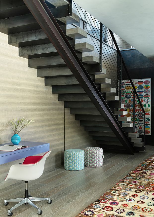 Interior view of a contemporary industrial-style stair hall and home office nook. A floating open-riser staircase with dark steel stringers, concrete treads, and black metal railing rises along a textured light wall. Below and beside it is a compact desk with a modern white swivel chair, colourful patterned ottomans, a bright blue vessel, artwork, and a large multicoloured patterned rug. The space combines raw structural elements with playful furnishings and strong natural light.
