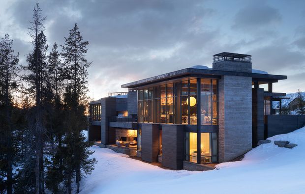A contemporary multi-level mountain house photographed at dusk, set among tall conifer trees and covered in snow. The residence has a strongly horizontal flat-roofed composition, extensive floor-to-ceiling glazing, dark metal or timber-look wall cladding, and a prominent grey masonry or concrete tower with a rooftop screened structure. Warm interior lighting glows through the windows, contrasting with the cool snowy landscape and overcast sky.