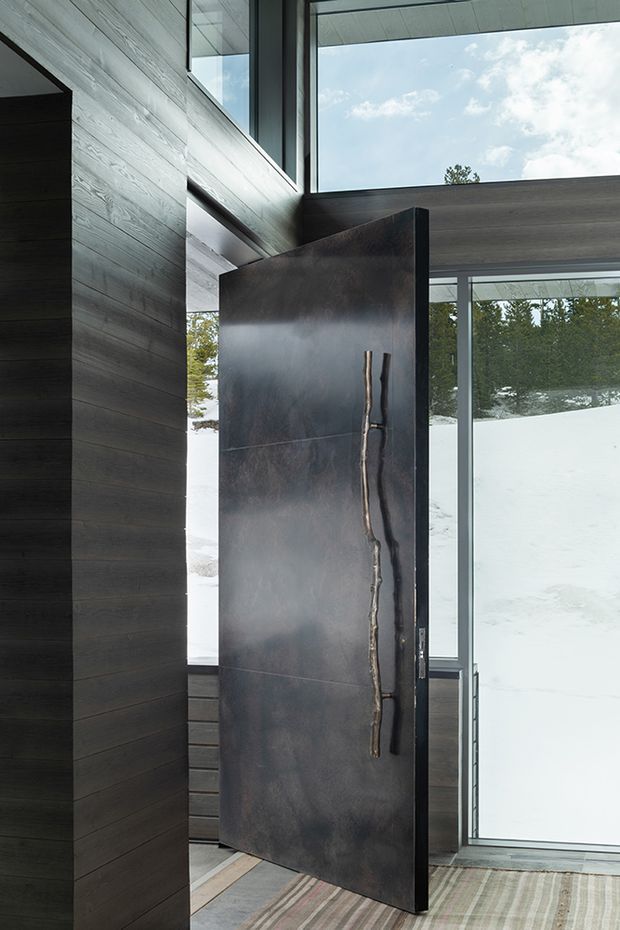 A dramatic contemporary entryway with a tall pivot front door in a dark, mottled metal finish and an elongated sculptural vertical handle. The entry is framed by dark horizontal timber wall panelling, exposed structural or ceiling elements, and large floor-to-ceiling glazing. Snow-covered terrain and trees are visible outside, while a muted striped rug softens the interior.