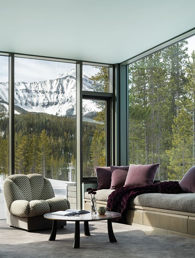A refined contemporary living room with floor-to-ceiling corner glazing overlooking a snow-covered mountain and dense alpine forest. The space features a built-in window seat with a grey upholstered cushion, deep plum cushions and throw, a textured cream lounge chair, a round dark-legged coffee table, and a pale grey carpet. Dark window frames and clean minimal surfaces create a calm, high-end mountain retreat atmosphere.