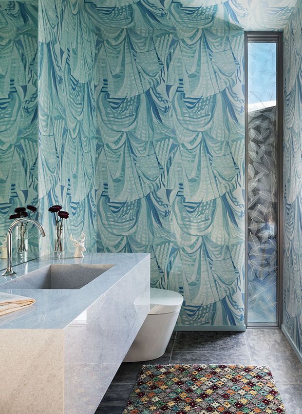 A compact, highly decorative bathroom featuring a freestanding-style integrated bathtub and vanity in pale stone, a wall-mounted or concealed toilet, and a narrow glazed shower enclosure. The walls are covered in bold blue-green patterned wallpaper, contrasted with dark grey stone-look flooring, a geometric patterned rug, brushed metal tapware, and a tall dark-framed window or glazed panel. The overall design is colourful, graphic, and refined.