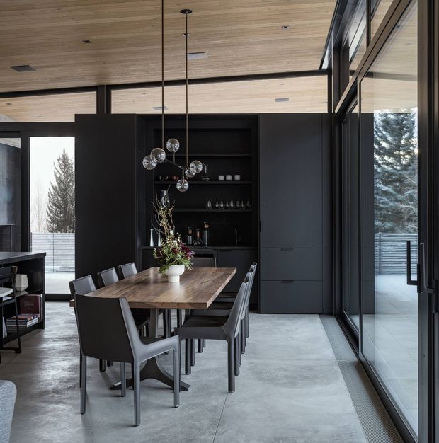 A sophisticated open-plan dining area with a substantial timber dining table and dark upholstered dining chairs. The room features a dark built-in bar and storage wall with open shelving, a sculptural multi-globe pendant light, expansive black-framed glazing, a pale concrete floor, and a warm timber-lined ceiling. The composition has a refined contemporary character with strong indoor-outdoor connection through the floor-to-ceiling windows.