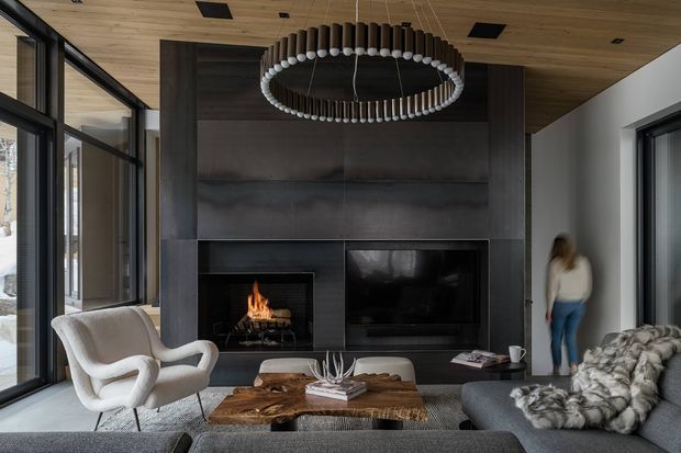 A sophisticated modern living room with a dramatic dark fireplace and integrated television wall, floor-to-ceiling glazing, a warm timber ceiling, and a large circular pendant light. Soft upholstered seating surrounds a substantial live-edge timber coffee table, while a lit fireplace adds warmth to the otherwise dark, refined palette.