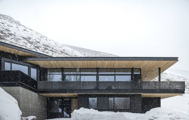 A modern, multi-level mountain residence photographed during heavy snowfall. The dark horizontal facade combines broad floor-to-ceiling glazing with projecting flat roof planes, deep timber-toned soffits, and expansive balconies or terraces. Snow-covered alpine slopes form the immediate setting.
