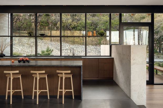 A refined contemporary kitchen with a large island and three timber bar stools, framed by expansive black-framed glazing overlooking a stone retaining wall and wooded landscape. Dark timber cabinetry, a pale stone benchtop, concrete-toned flooring, and a sculptural plaster or concrete partition create a warm yet minimalist indoor-outdoor composition.