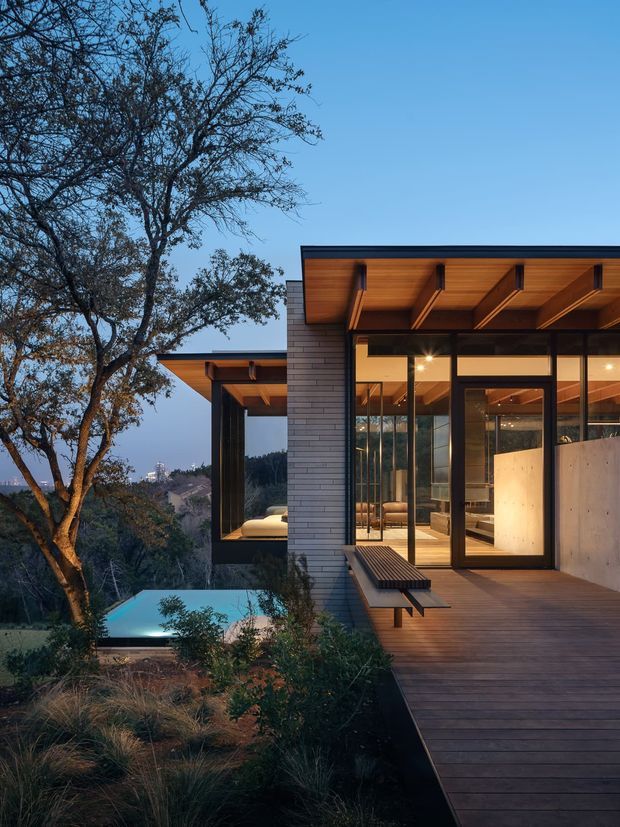 Exterior architectural photograph of a contemporary hillside home at dusk. The composition highlights dark horizontal cladding, extensive floor-to-ceiling glazing, warm timber soffits with exposed projecting beams, and a broad timber deck with built-in bench seating. A swimming pool and landscaped native planting sit below the house, with mature trees and a distant wooded landscape beyond.